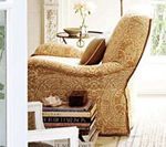 Cozy armchair with patterned gold fabric, a small side table with a vase, and a stack of books.
