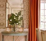 A vase of white flowers on a blue console table, reflected in a large mirror. Orange curtains.
