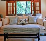 A living room with a cream-colored couch, patterned ottoman, and a large window looking outside.