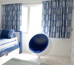 Blue and white patterned curtains frame a sunny window, next to a matching accent chair and bed.