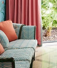 Sofa with blue patterned cushions, peach pillow, and pink curtains next to a window with green plants.
