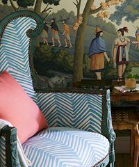 Upholstered chair with blue and white zigzag fabric, pink pillow, against mural with figures in a landscape.