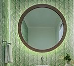 Round mirror with wooden frame on green leaf-patterned wall, above a sink.