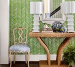 A console table with lamps and decor against a green patterned wall, with a chair nearby.