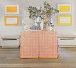 A room with peach-colored cabinet, white chests, yellow framed artwork, and decorative objects.