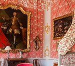 Ornate room with red patterned wallpaper, large gold-framed portraits, and floral-draped bed.