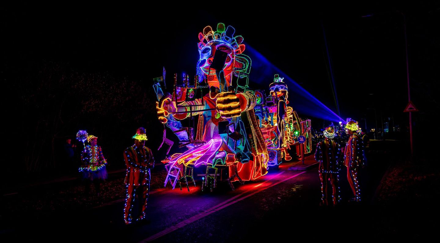 Een praalwagen in de avond met felle neonlichten en artiesten in verlichte kostuums.