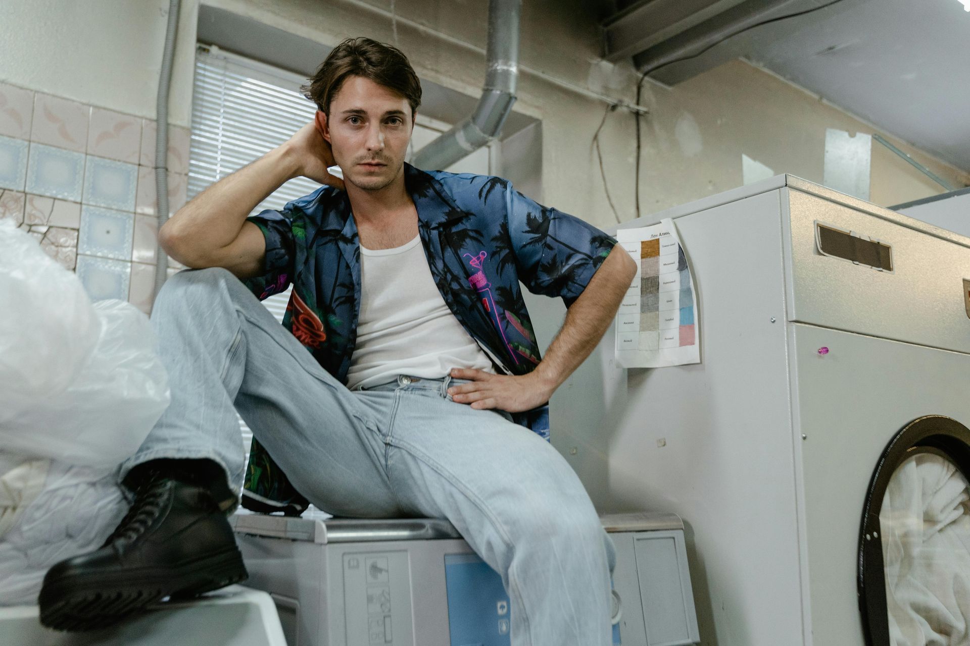 A man is sitting on top of a washing machine in a laundromat.