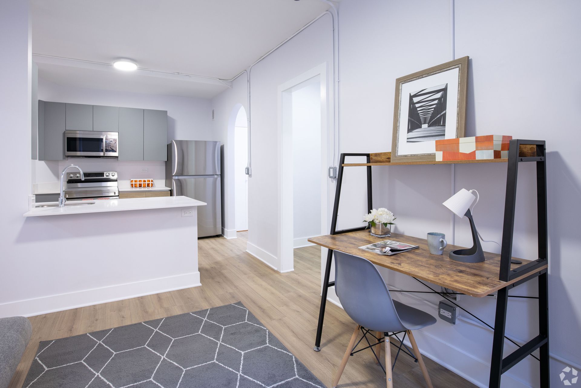 A room with a desk and chair in it and a kitchen in the background.