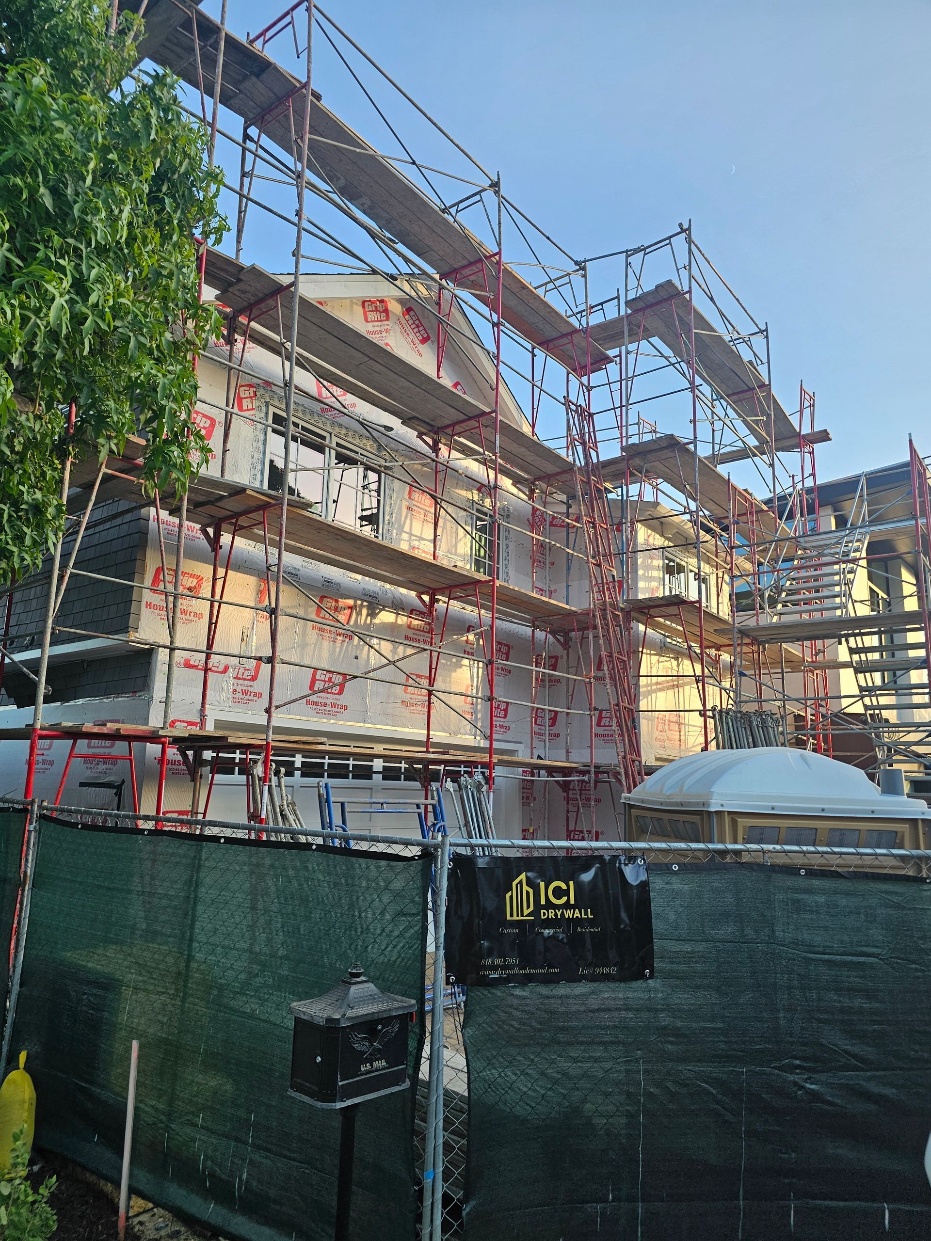 Scaffolding around a multi-story building under construction behind a green fence, with blue sky above