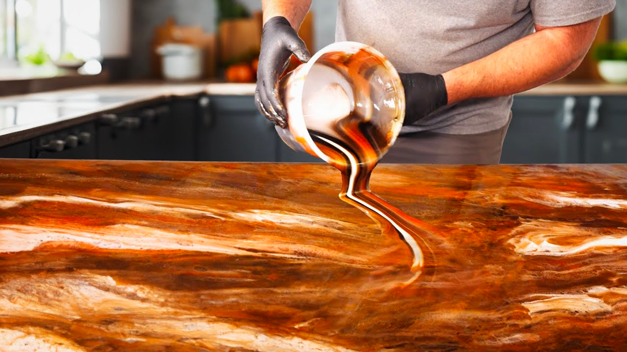 A person pours a dark, swirling liquid onto a wood countertop. The liquid creates a marbled pattern.