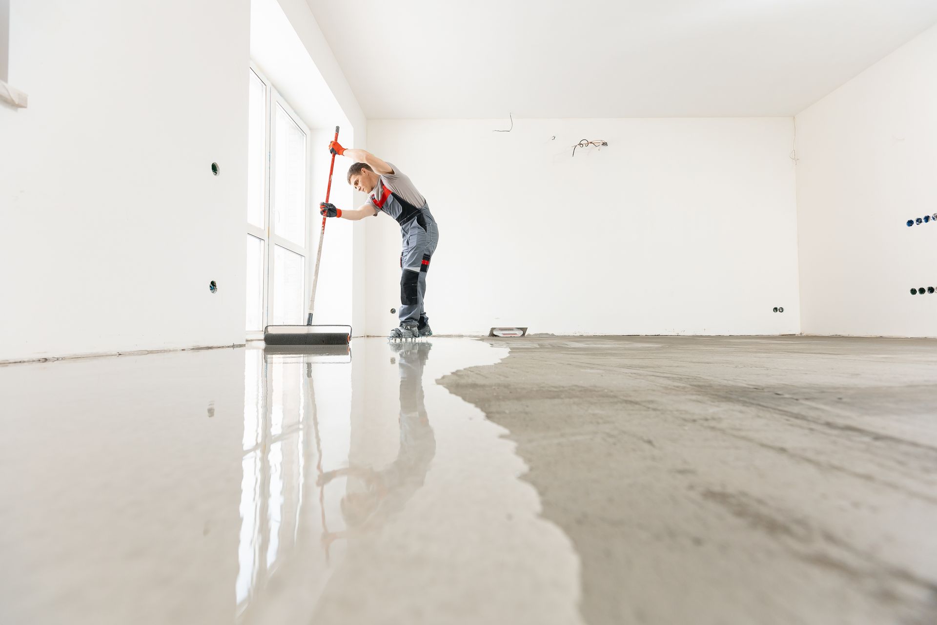 Person leveling wet concrete floor in a room with white walls, using a large squeegee.