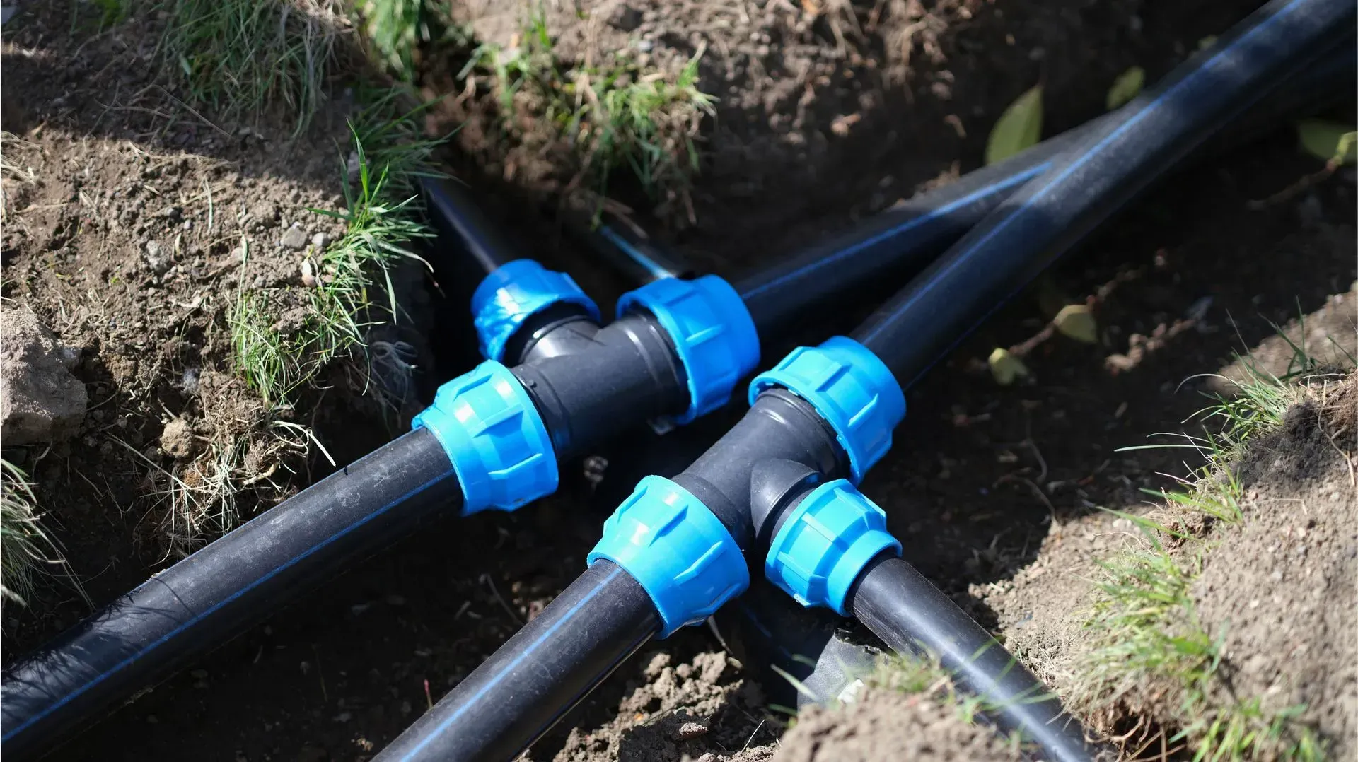 Black plastic pipes connected by blue compression fittings, arranged in a T-junction layout in a dirt trench.