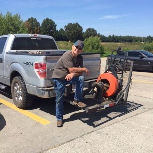 A person posing next to a plumbing drain snake machine mounted on a truck hitch in a parking lot.
