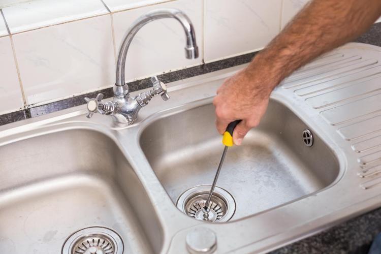 A person uses a screwdriver to repair a metal kitchen sink drain.