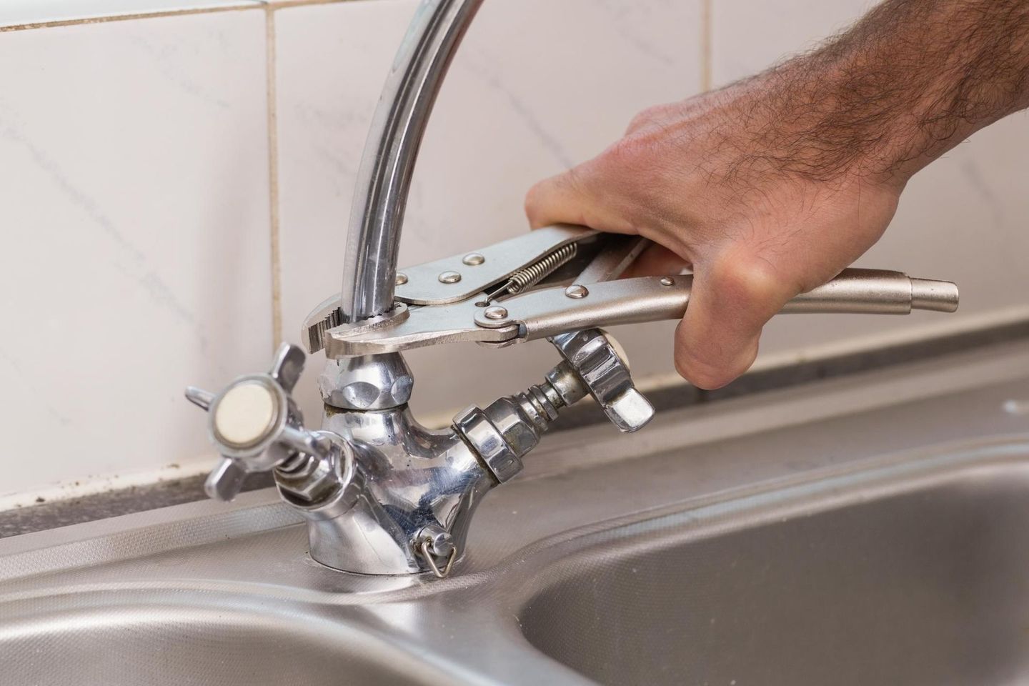 A person uses locking pliers to grip the neck of a chrome kitchen faucet above a stainless steel sink.