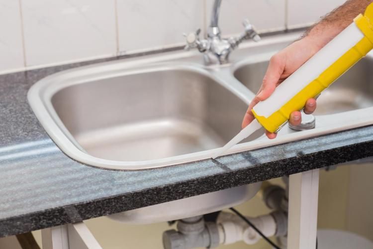 A hand uses a yellow caulk gun to apply sealant along the edge of a stainless steel kitchen sink and granite countertop.
