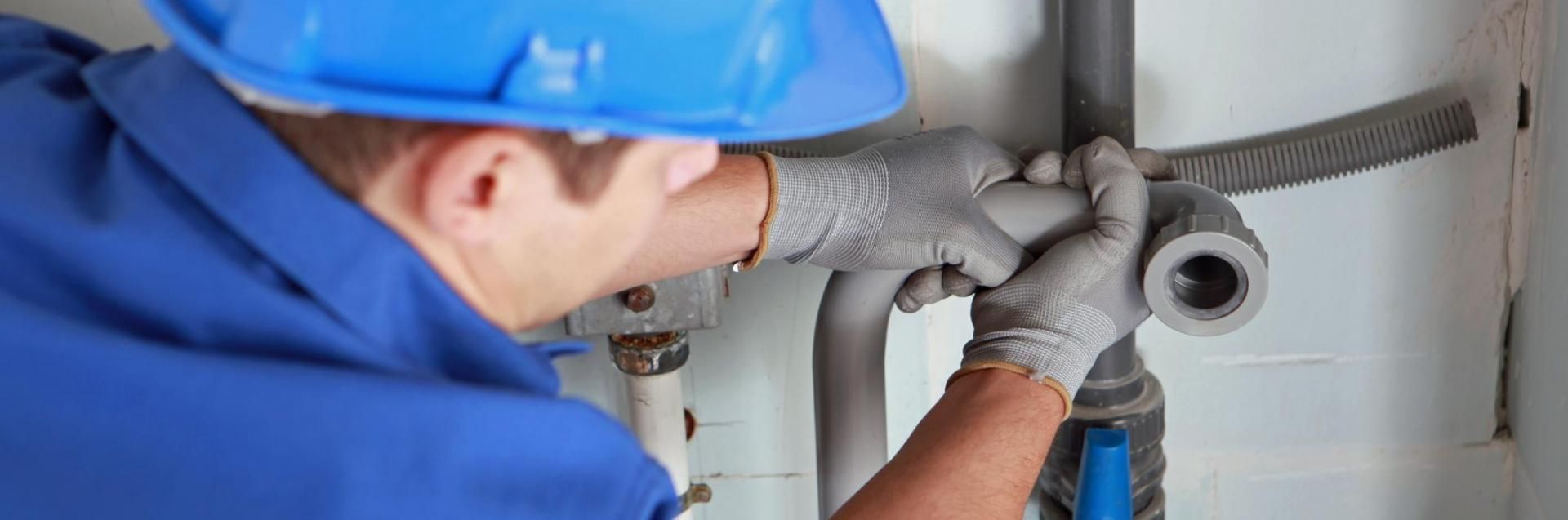 A person in a blue hard hat and uniform wears gray gloves while working on gray plumbing pipes against a white wall.