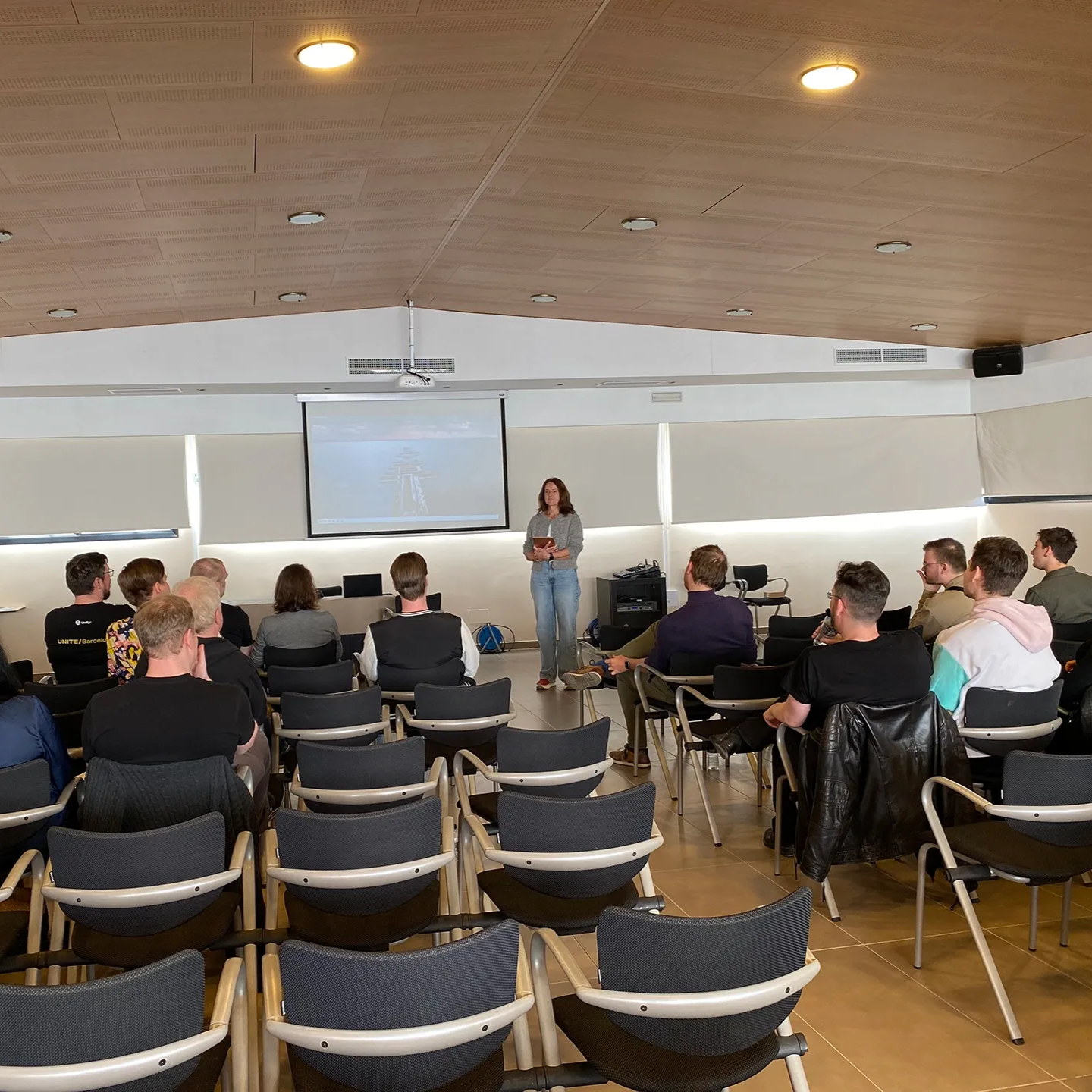 Eine Rednerin steht vor einer Projektionsfläche in einem modernen Konferenzraum und spricht zu einem Publikum sitzender Personen.