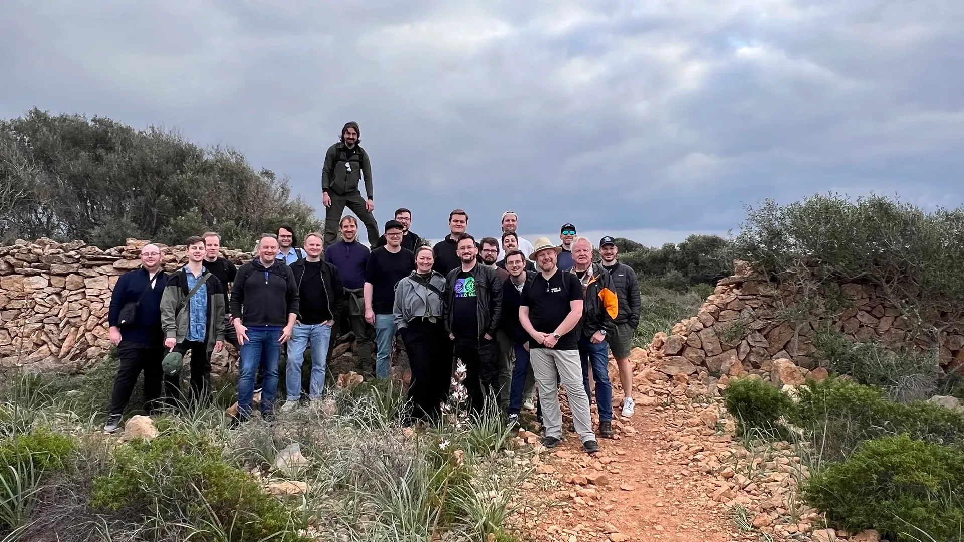 Eine Gruppe von Menschen steht an einem bewölkten Tag im Freien auf einem unbefestigten Pfad in der Nähe von Steinmauern.