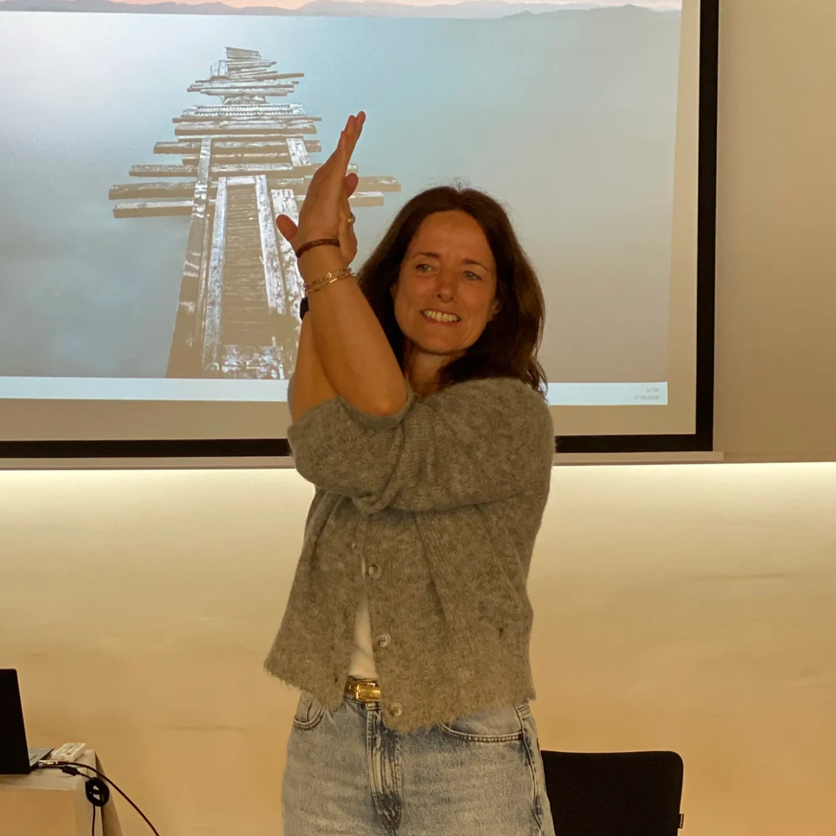 Eine Frau in grauer Strickjacke und Jeans führt eine Armbalance-Yoga-Pose vor der Projektion eines Holzstegs aus.