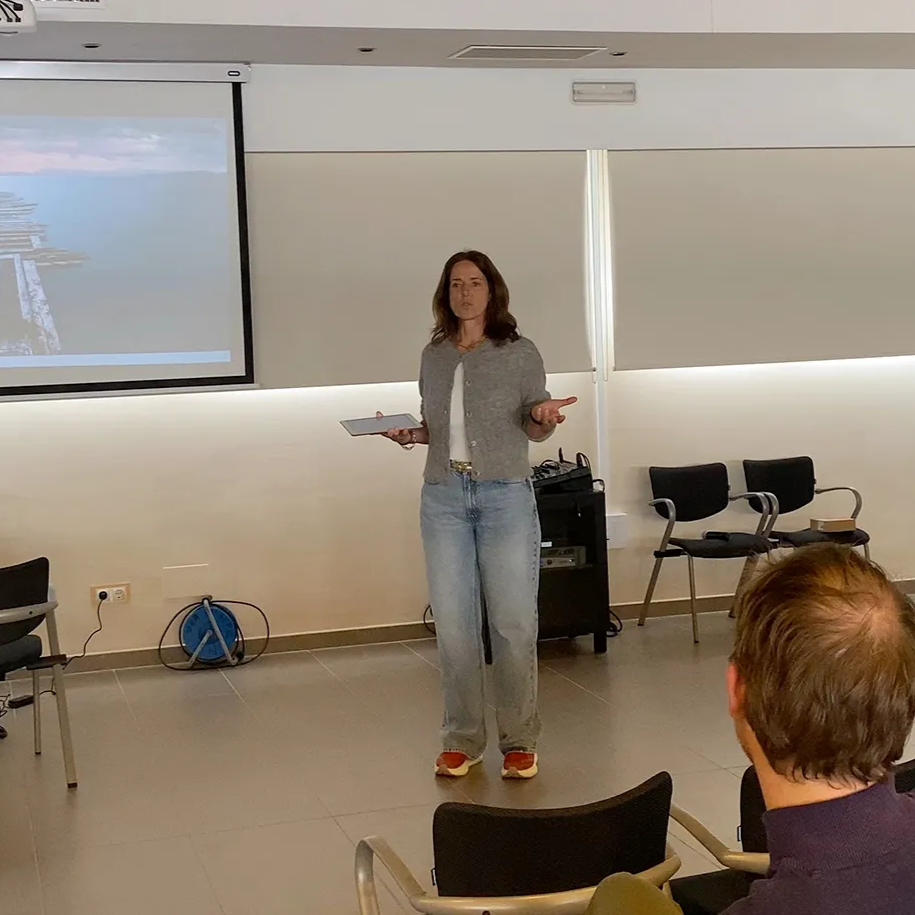 Eine Moderatorin steht in einem Raum mit einer Projektionsfläche und gestikuliert, während sie zu einem Publikum spricht.