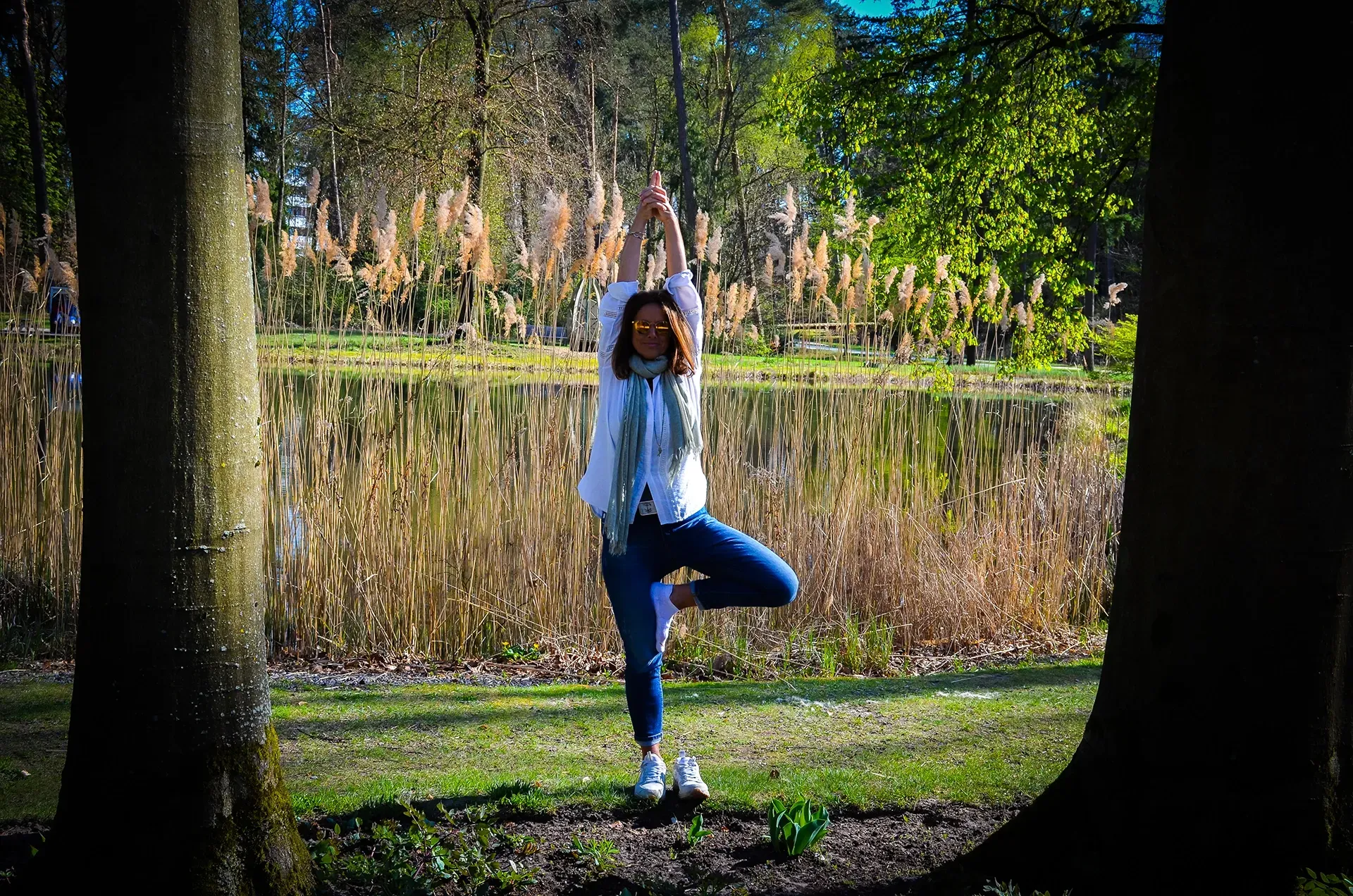 Eine Frau steht in einer Baum-Yoga-Pose, die Arme erhoben, auf einer Rasenfläche neben einem von Bäumen umgebenen Teich.