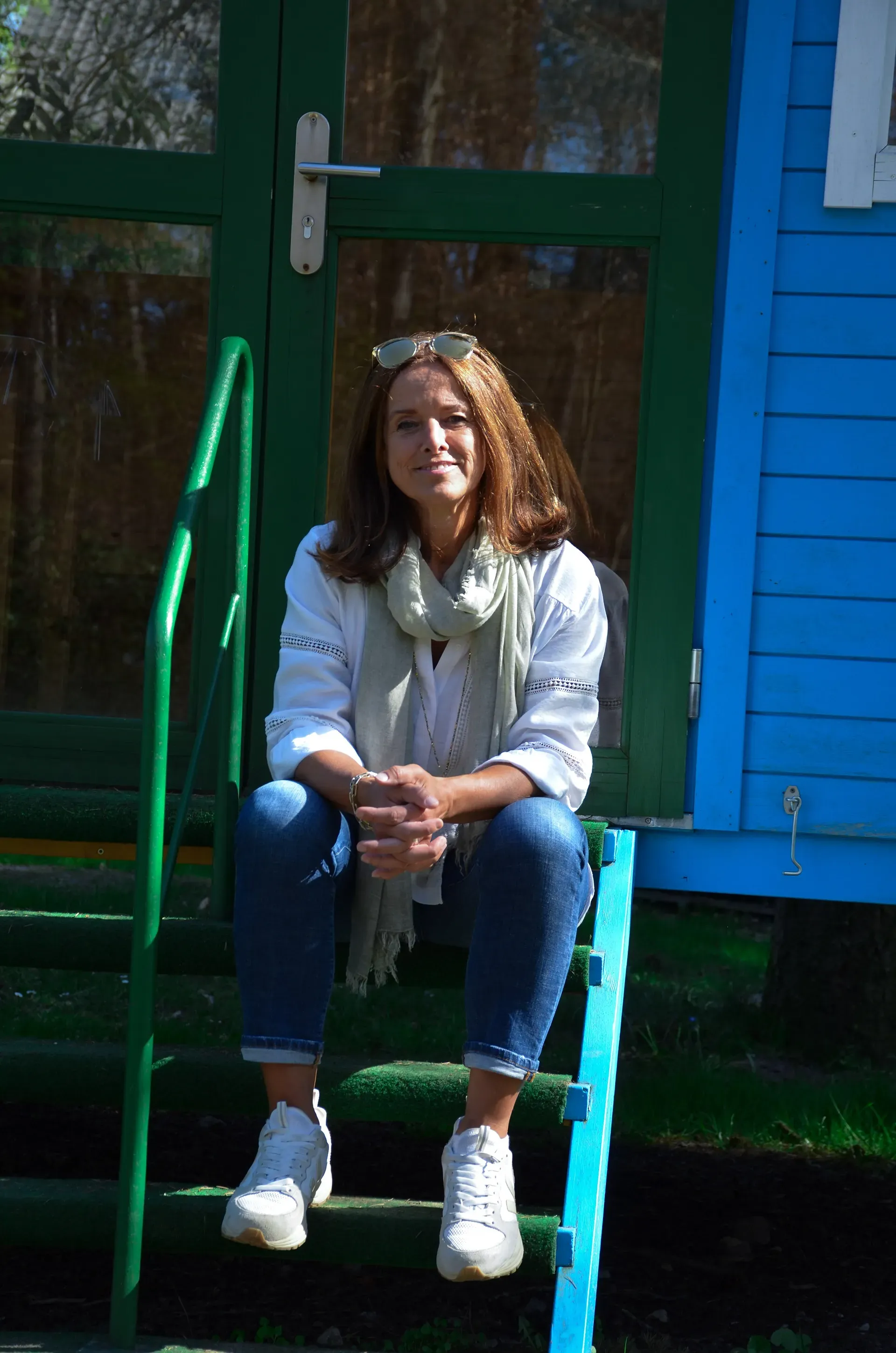 Eine lächelnde Frau sitzt auf einer grünen Außentreppe vor einem blauen Gebäude. Sie trägt ein weißes Hemd, Jeans und einen Schal.