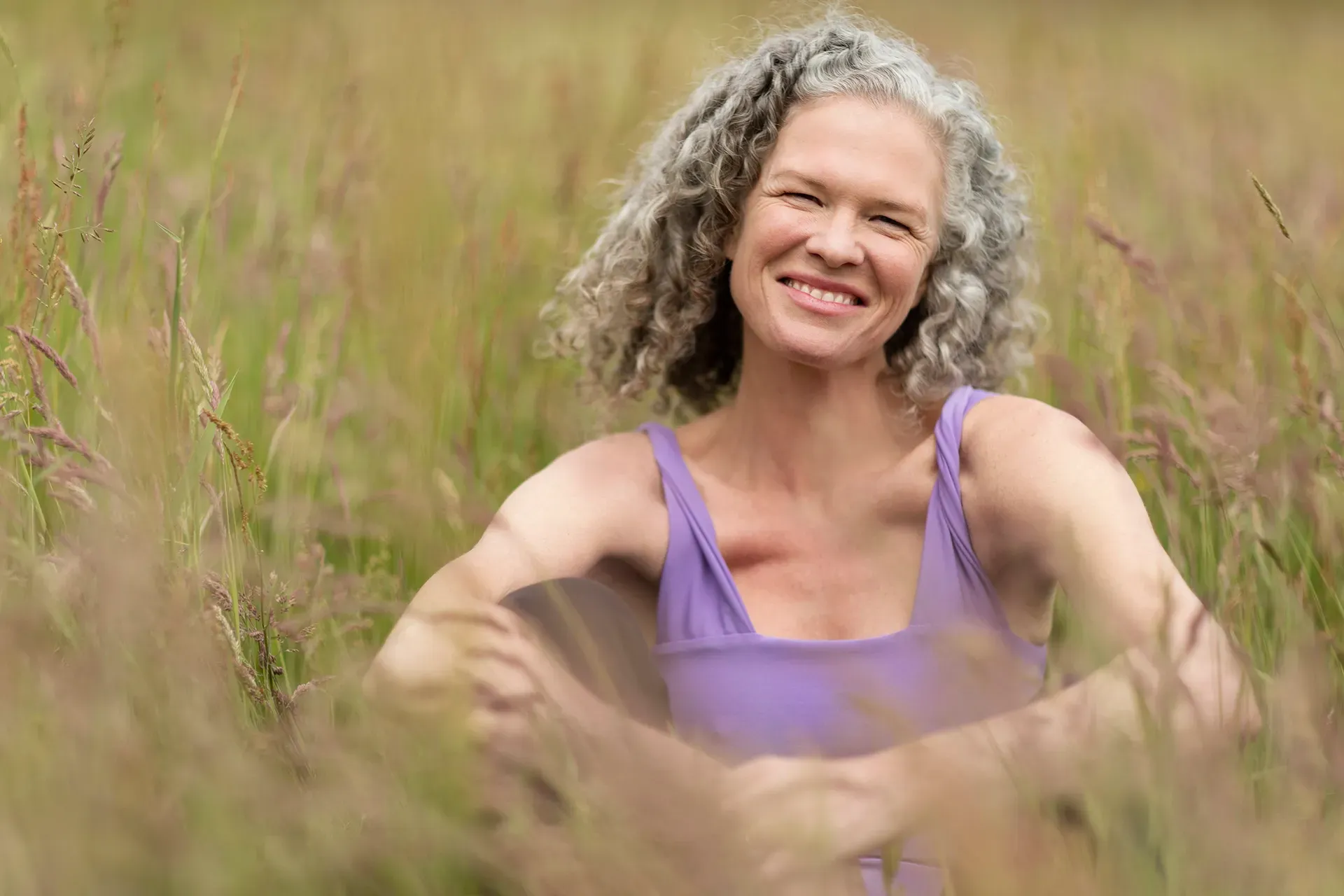 Eine lächelnde Person mit lockigem Haar, die ein lila Tanktop trägt, sitzt in einem Feld mit hohem Gras.