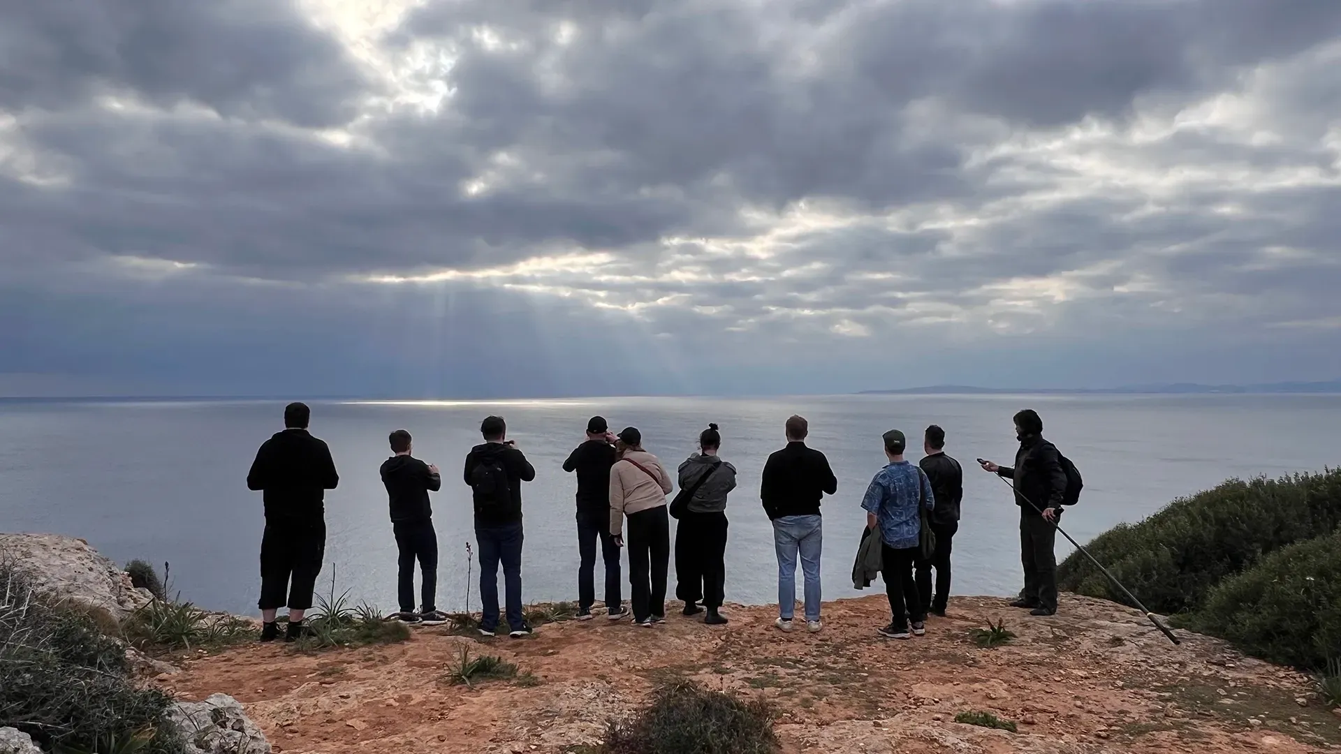 Eine Gruppe von Menschen steht auf einer felsigen Klippe und blickt unter einem bewölkten Himmel auf einen ruhigen, weiten Ozean hinaus.
