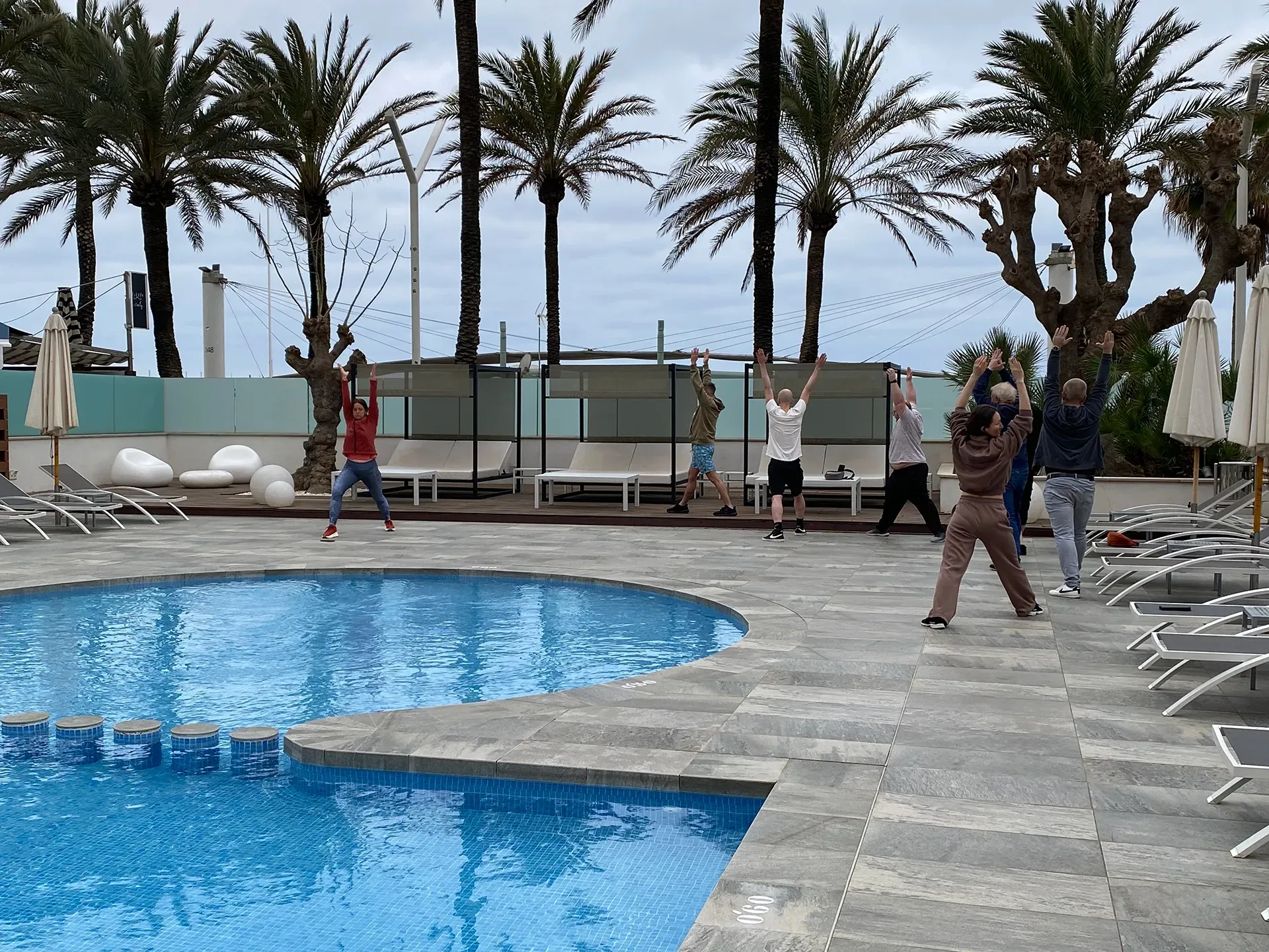 Eine Gruppe von Menschen steht mit erhobenen Armen auf einer steinernen Poolterrasse während eines Gruppenyoogakurses.