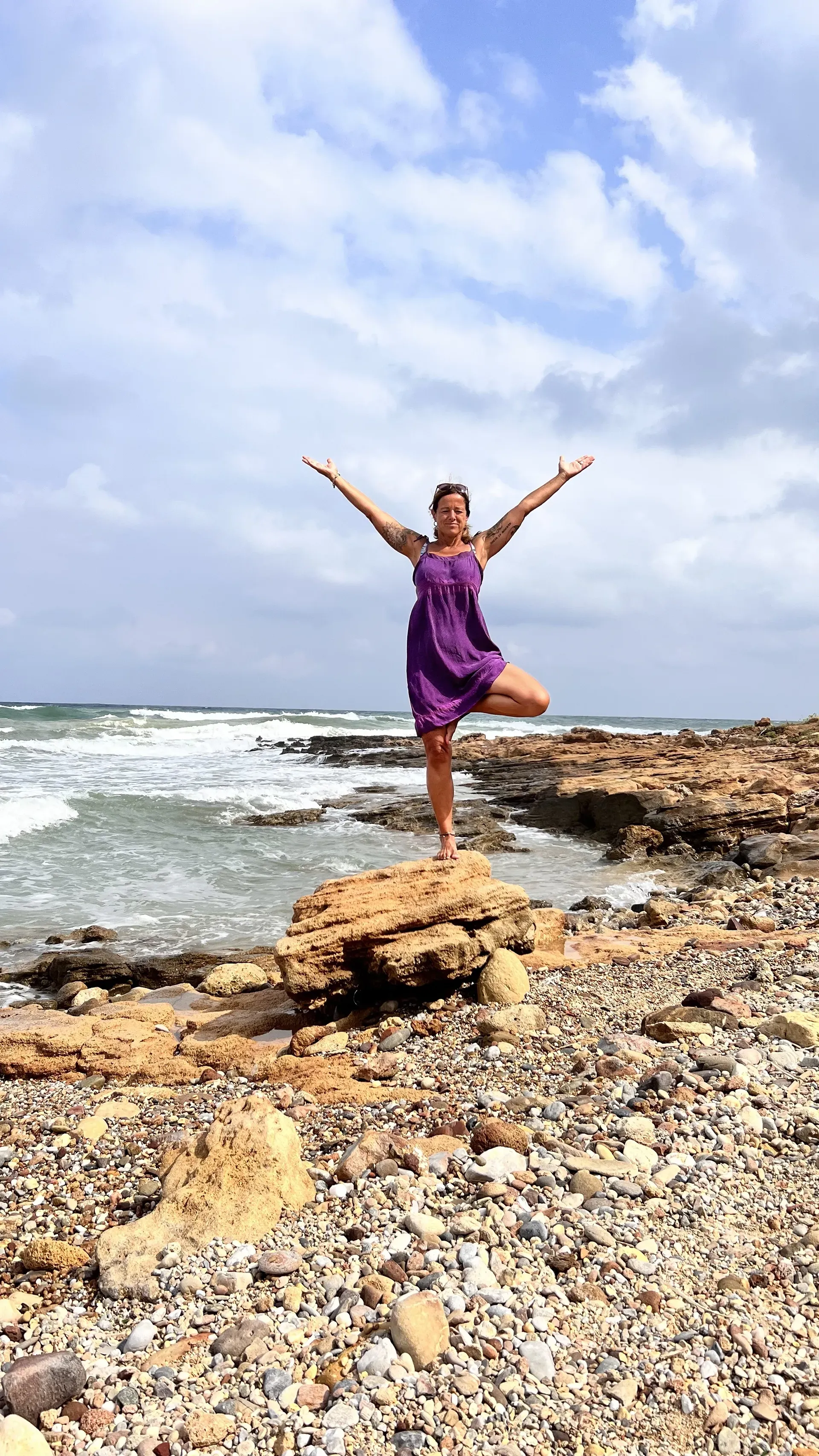 Eine Frau in einem lila Kleid balanciert in einer Baumpose auf einem felsigen Strand am Meer unter einem bewölkten Himmel.