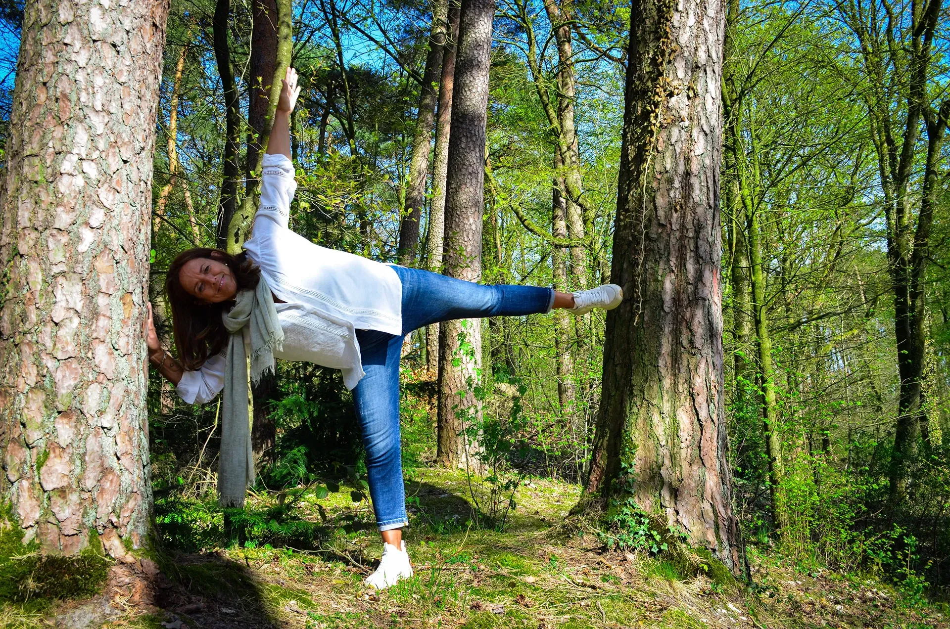 Eine Frau balanciert auf einem Bein in der Seitstützposition, den Arm nach oben ausgestreckt, zwischen zwei Bäumen in einem sonnigen Wald.