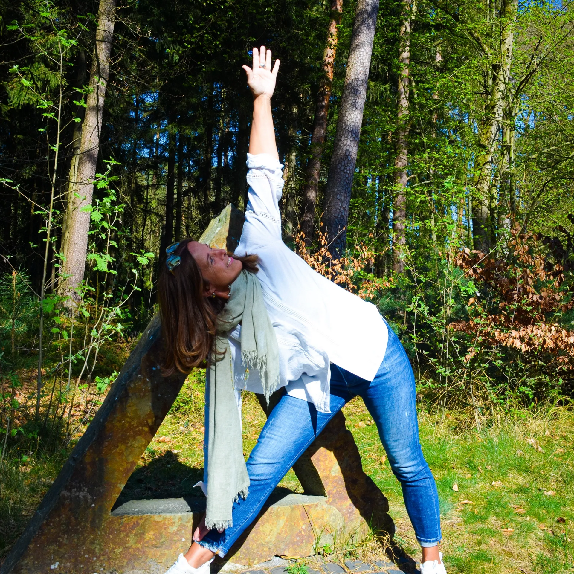 Eine Frau in Jeans und weißem Hemd führt in einem sonnigen, bewaldeten Außenbereich die Yoga-Dreieckspose aus.