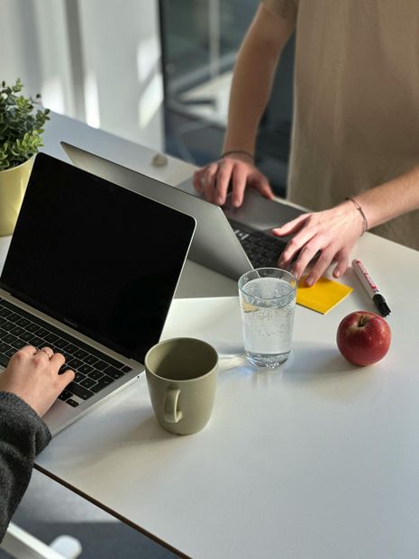 Zwei Personen arbeiten an getrennten Laptops an einem weißen Schreibtisch. Neben ihnen stehen eine Tasse, ein Glas Wasser, ein gelber Zettel und ein roter Apfel.