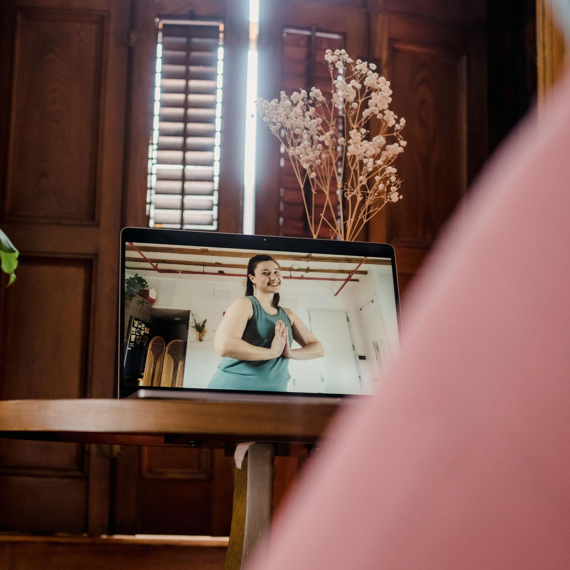 Eine Person trainiert in einem Online-Video, das auf dem Bildschirm eines Laptops angezeigt wird, der auf einem Holztisch vor Jalousien steht.