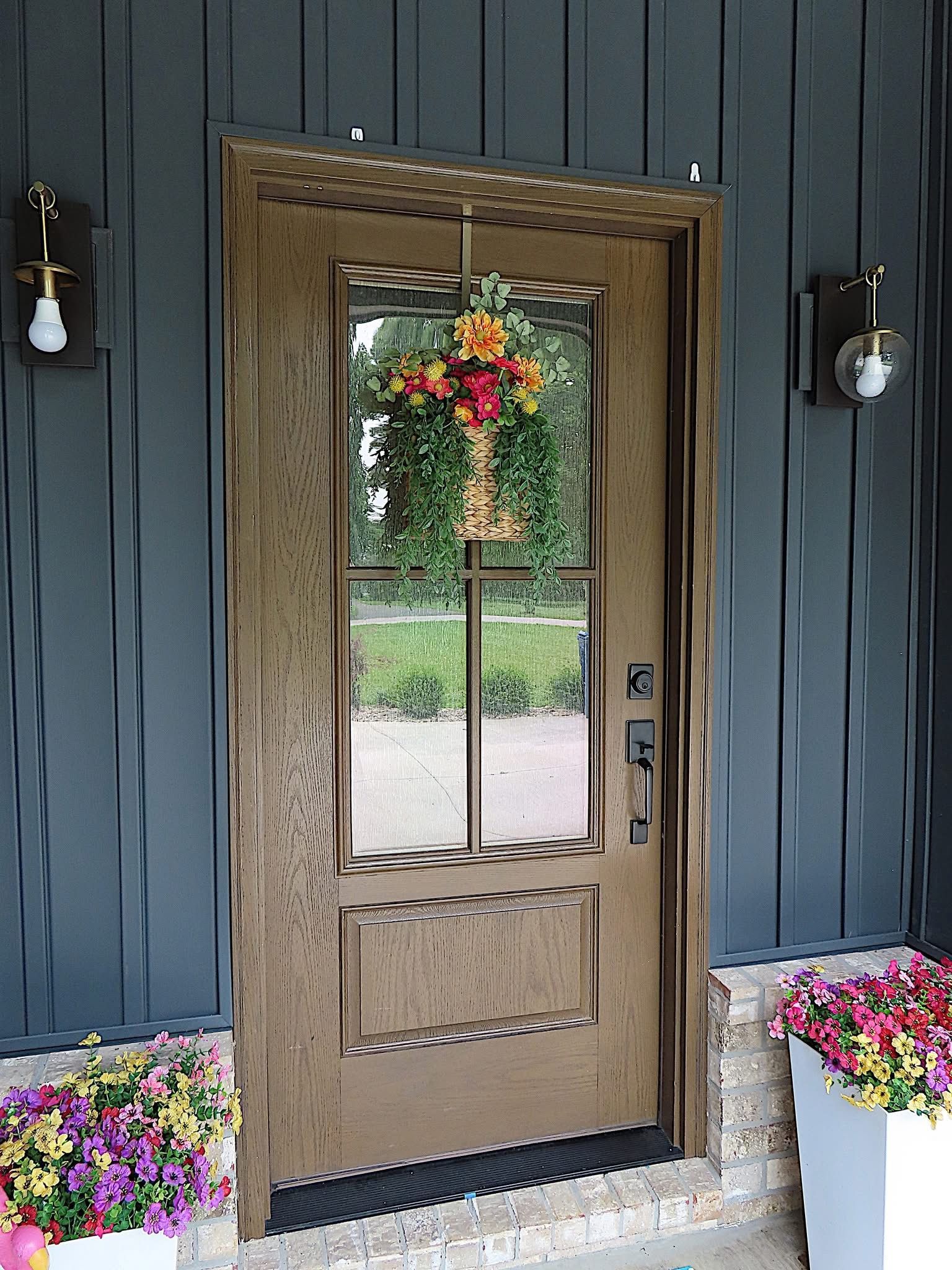 Brown front door with glass panel, floral wreath, flanked by outdoor lights, potted flowers.