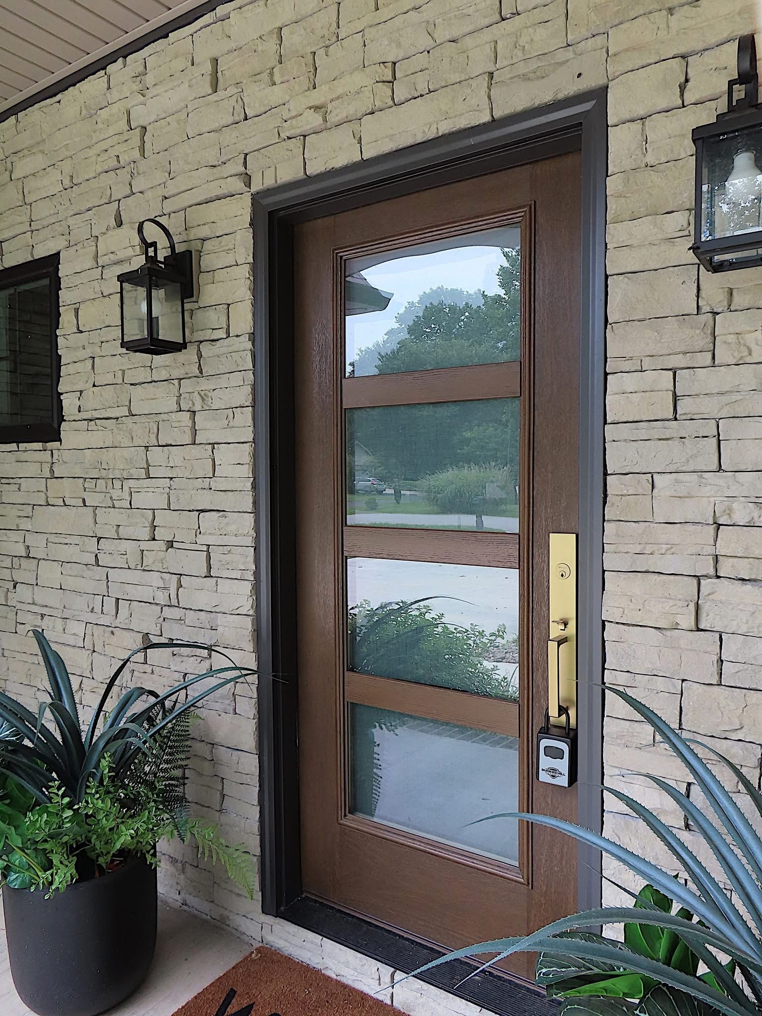 Brown front door with rectangular glass panes, dark metal trim, and gold hardware. Exterior with stone wall and plants.