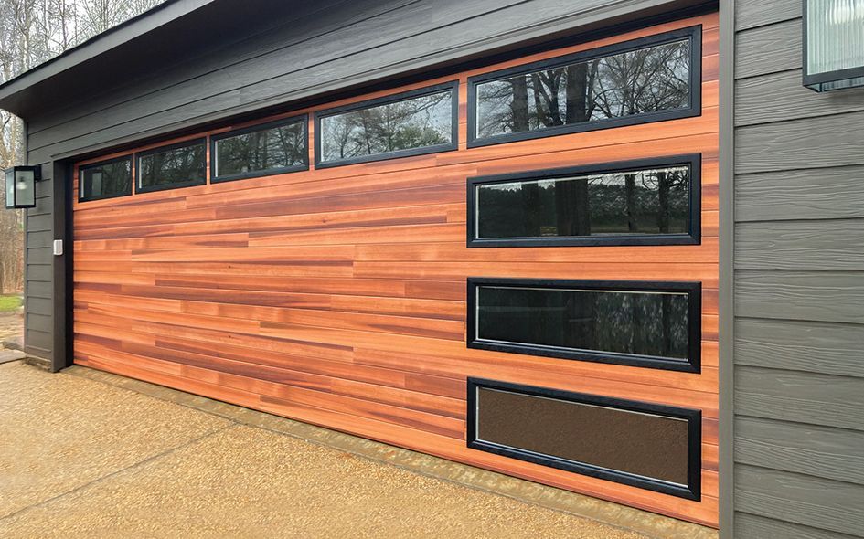 Wooden garage door with horizontal planks, black-framed windows, gray siding, and a concrete driveway.