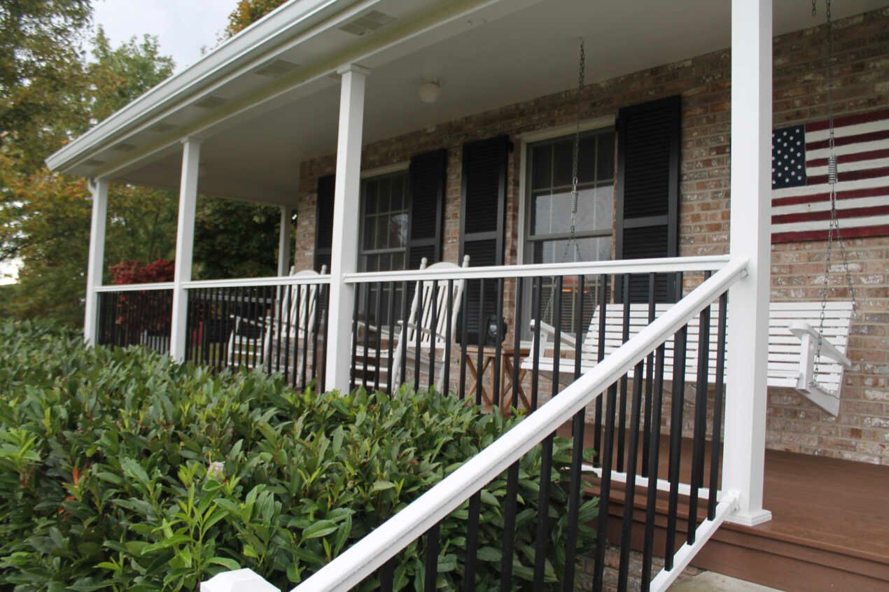 Front porch with white columns, black railings, and American flag. Green bushes in foreground.