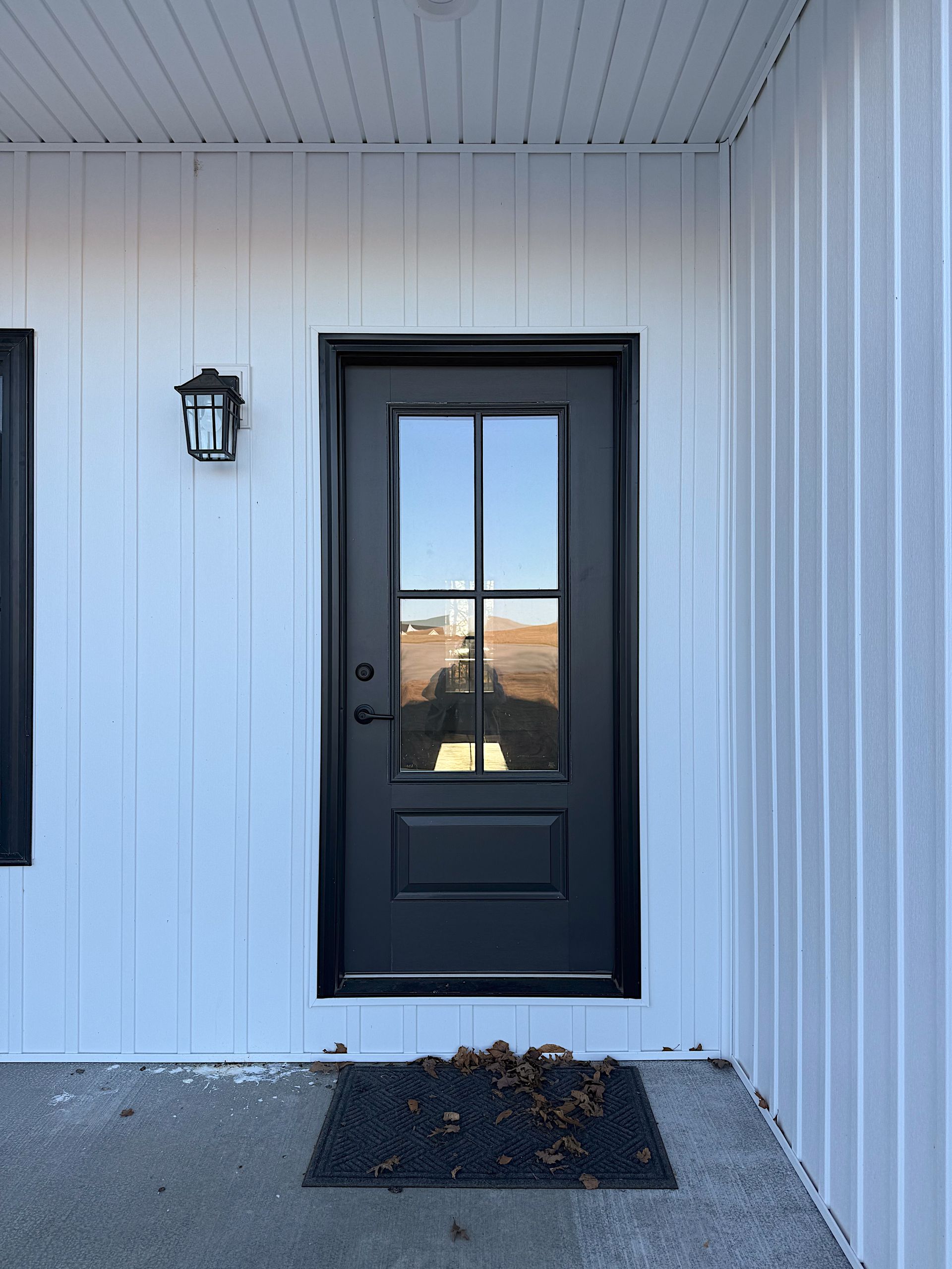 Black door with glass panes set in white siding; small black mat on concrete porch; black light fixture.