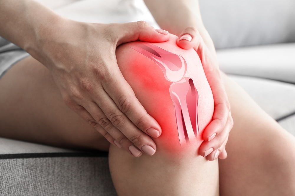 A Woman Is Sitting on A Couch Holding Her Knee in Pain — Illawarra Sports Injury Rehabilitation and Physiotherapy Centre in Bellambi, NSW