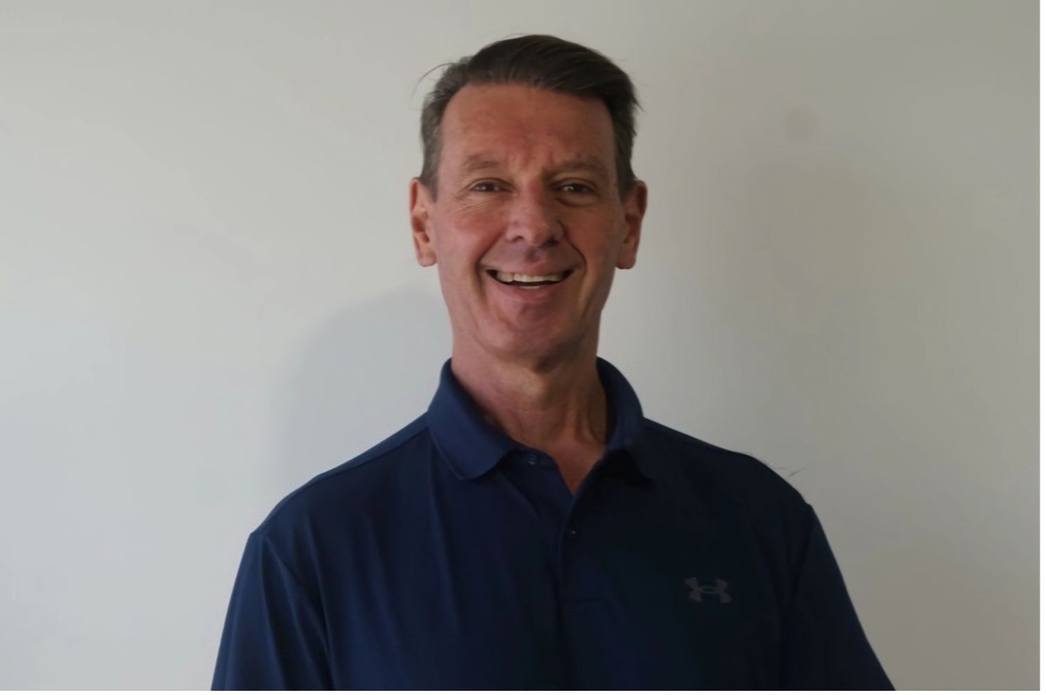 A Man Wearing a Blue Under Armour Shirt Smiles for The Camera — Illawarra Sports Injury Rehabilitation and Physiotherapy Centre in Bellambi, NSW