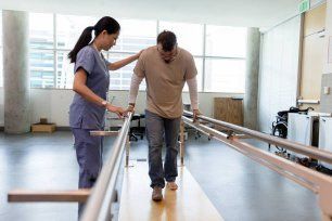 A Nurse Is Helping a Man Walk on Parallel Bars in A Hospital — Illawarra Sports Injury Rehabilitation and Physiotherapy Centre in Bellambi, NSW