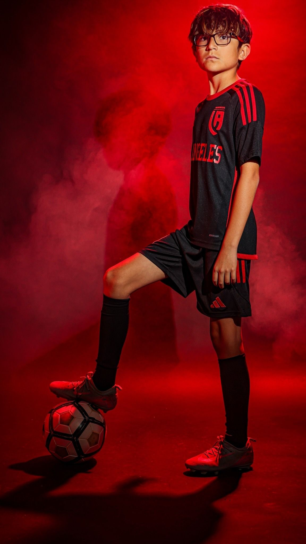 Soccer player in black and red uniform standing with one foot on a ball against a red background