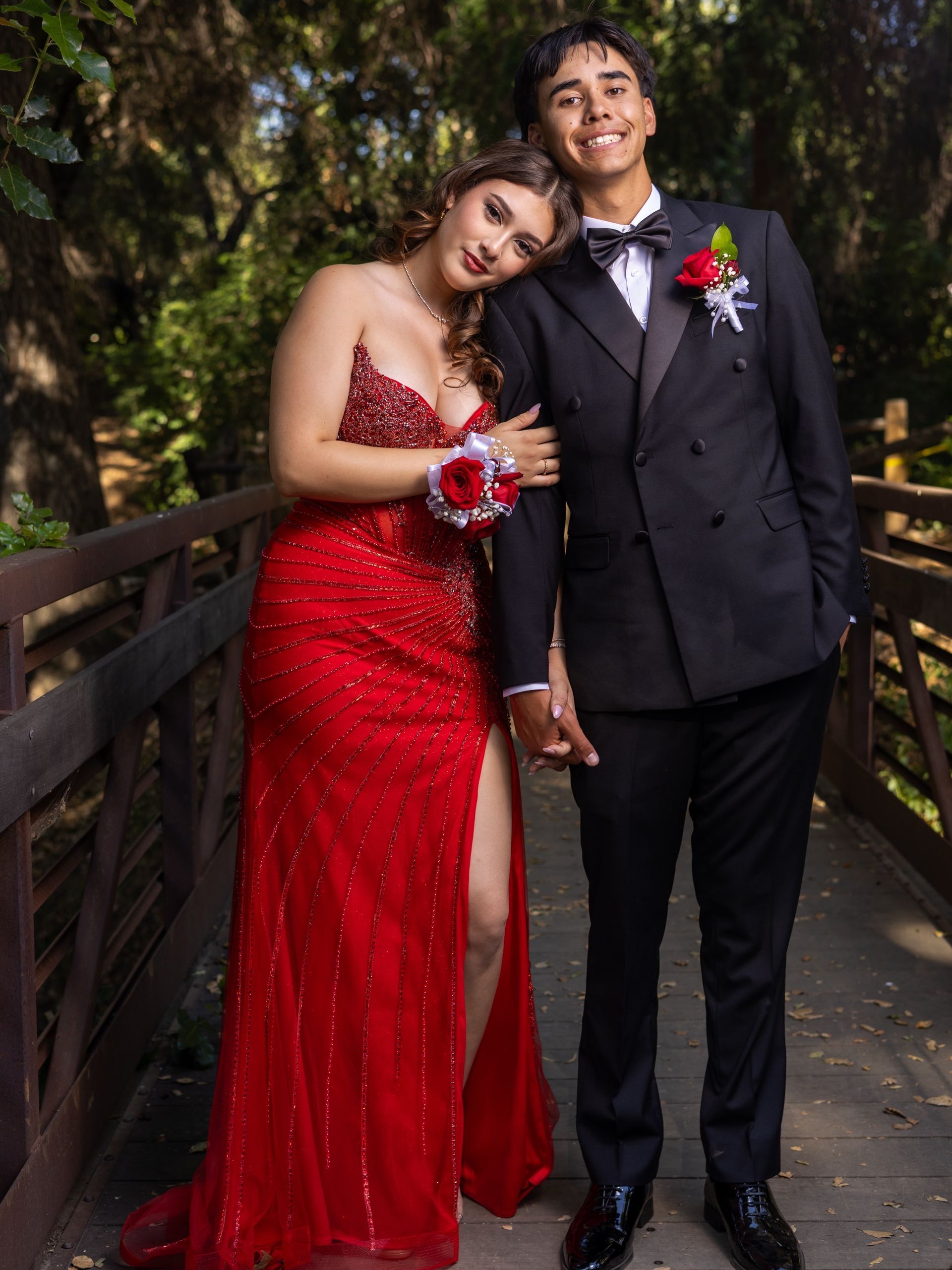 Couple in formal attire posing on a wooden bridge, woman in red dress leaning on man in black suit.