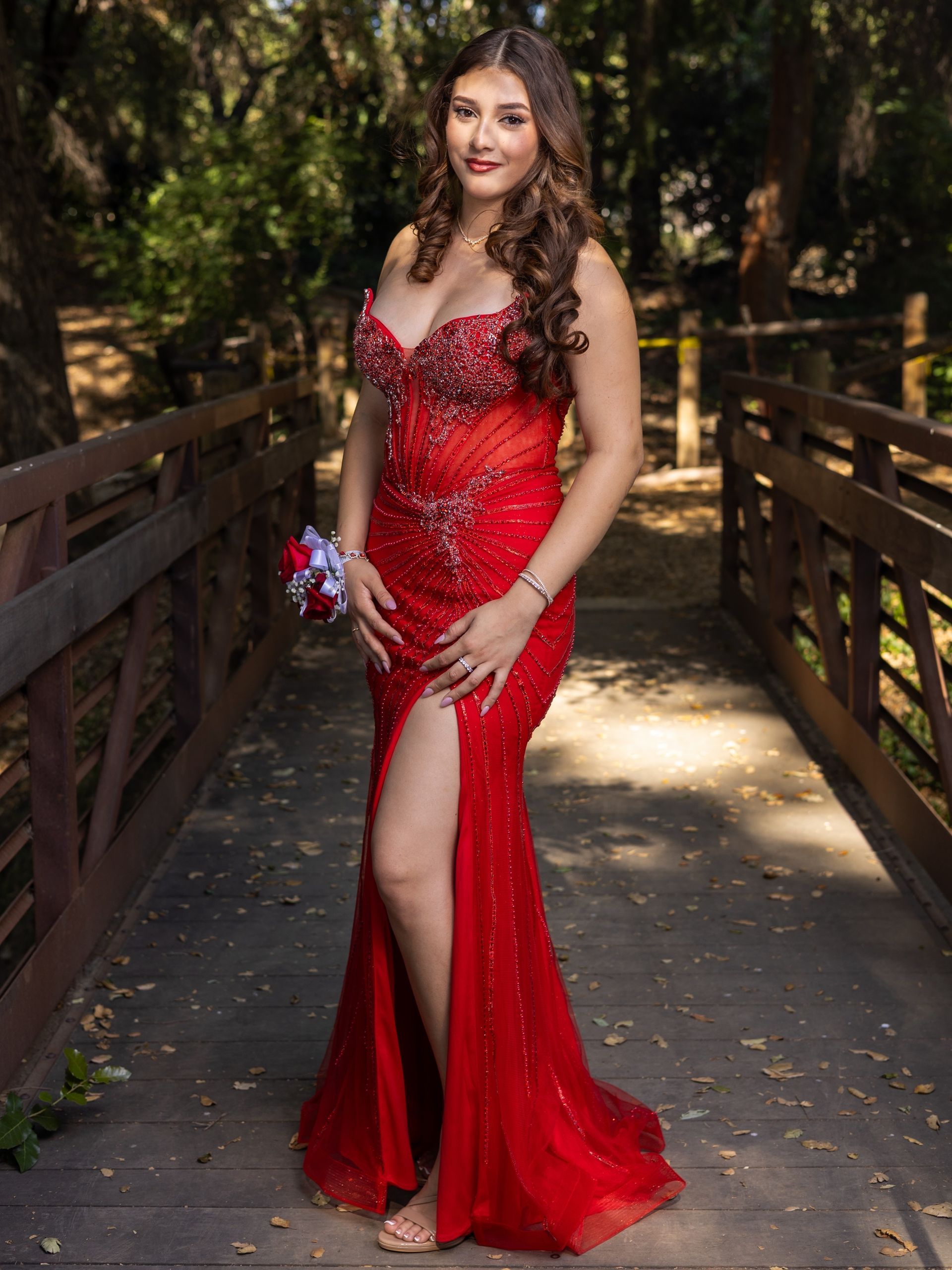Woman in a red sequined gown standing on a wooded bridge, holding a bouquet.