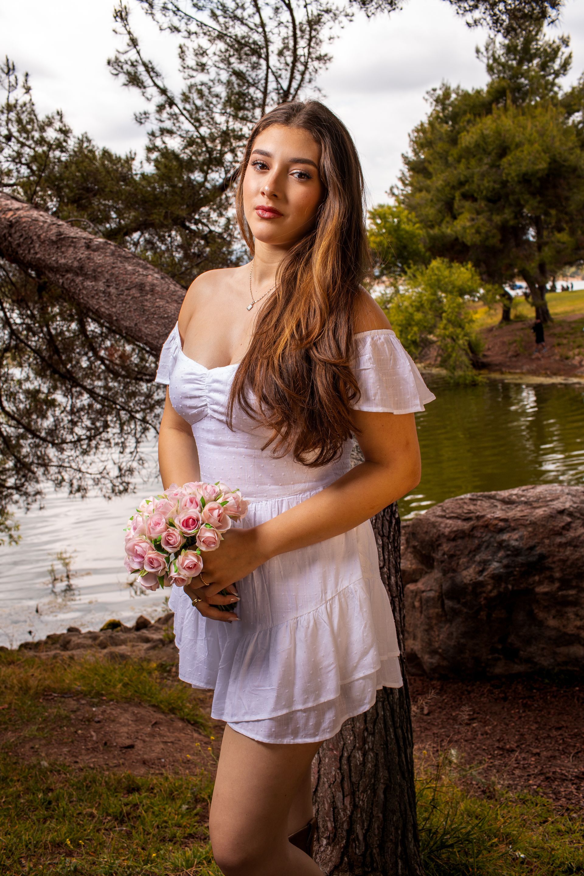 Woman in a white dress holding a pink bouquet by a tree beside a pond.
