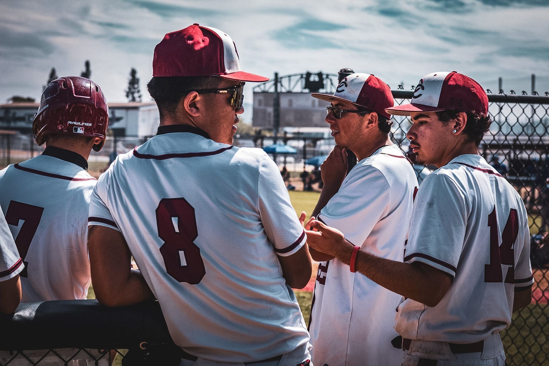 Baseball players in white uniforms and red caps huddle on a sunny field near the dugout.