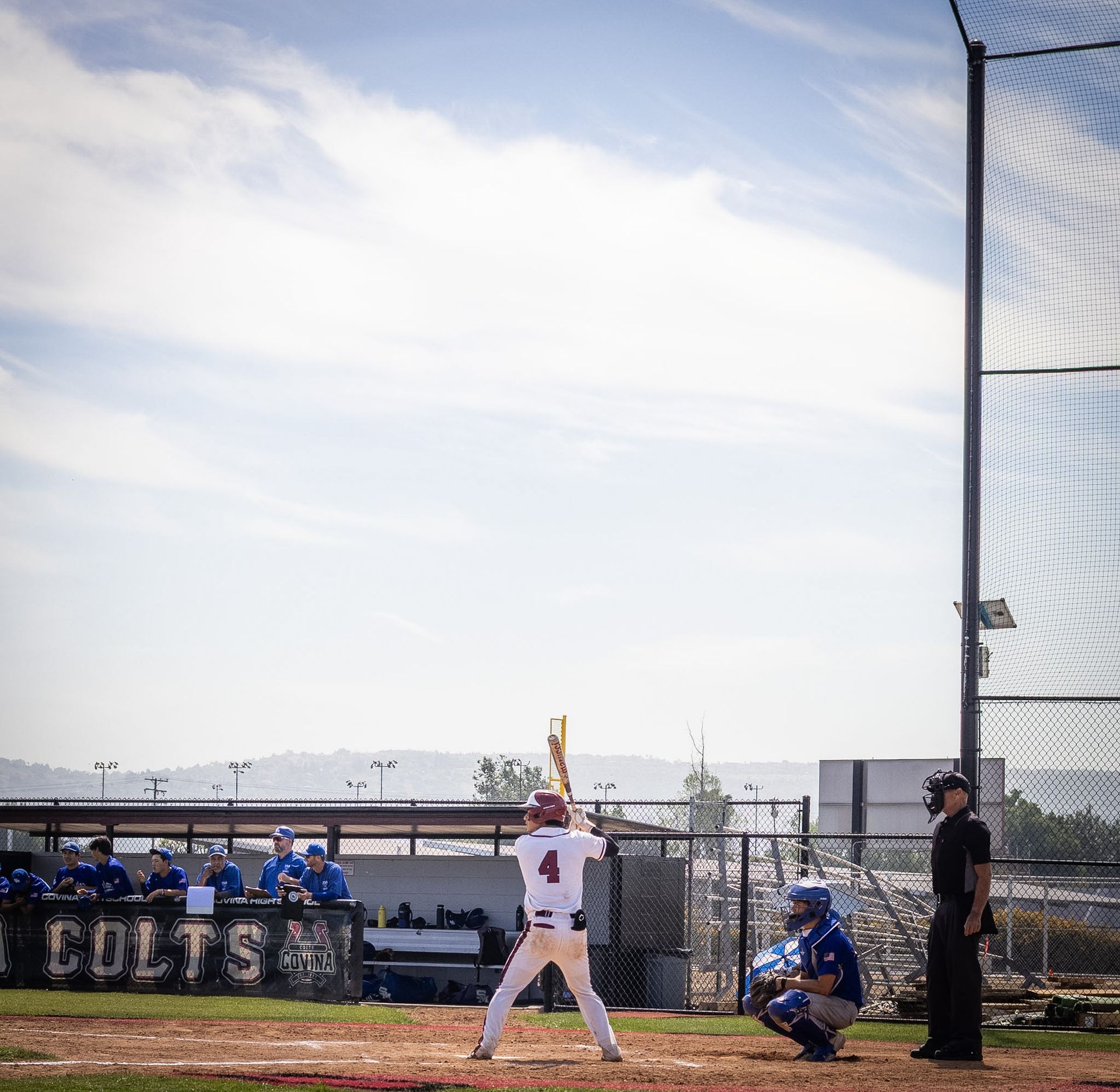 Baseball batter at home plate in a blue-and-white stadium, with catcher and umpire nearby under a bright sky